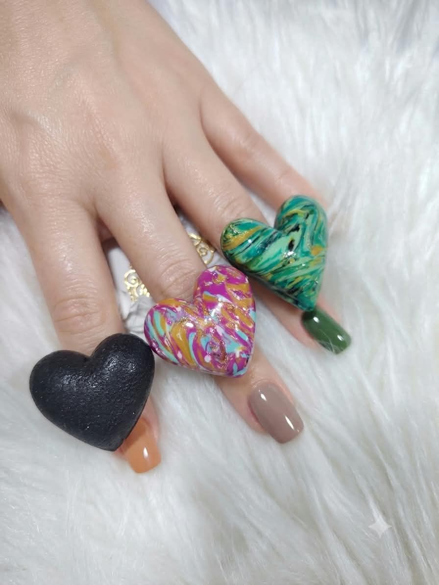Tres anillos de corazón de arcilla polimérica en una mano: negro, marmoleado fucsia y marmoleado verde